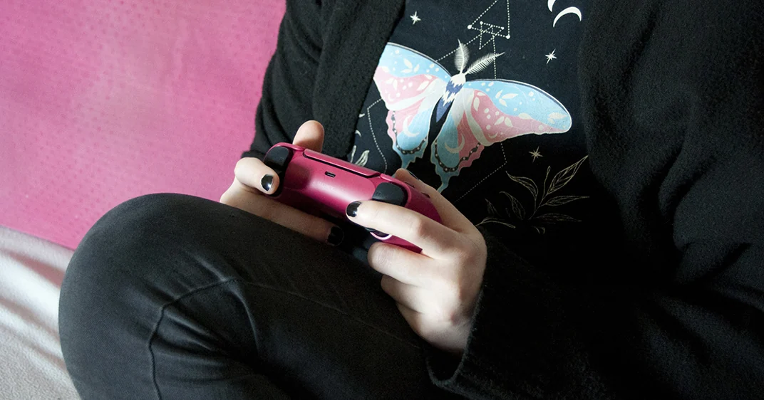 "A person sitting on a pink couch is holding a pink PlayStation 5 controller with both hands. Their body is oriented to the left. They are dressed in black, and their T-shirt is printed with a colorful butterfly. Their thumbs are on the ana